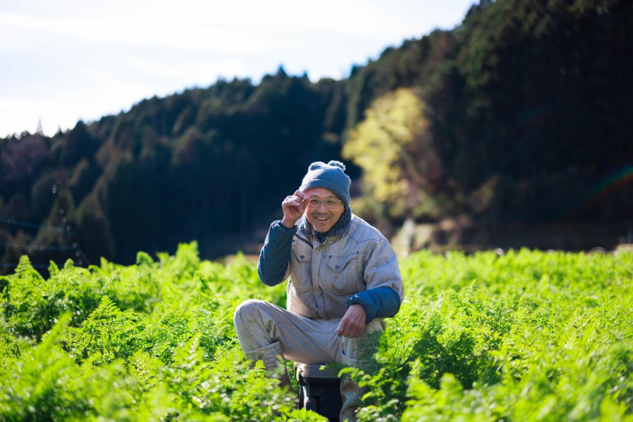 A smiling farmer in winter gear kneels in a lush field, representing the new generation revitalizing Japan’s farmlands.